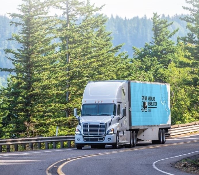Un camion-remorque bleu sur lequel est inscrit « Eye Van », circule sur une route sinueuse bordée d'arbres à feuilles persistantes.