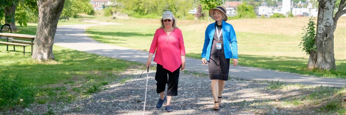 Two women walk down outdoor path, one uses white cane 