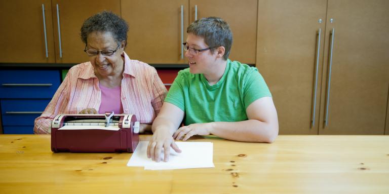 Une femme plus âgée et une femme plus jeune sont assises à une table devant une machine braille