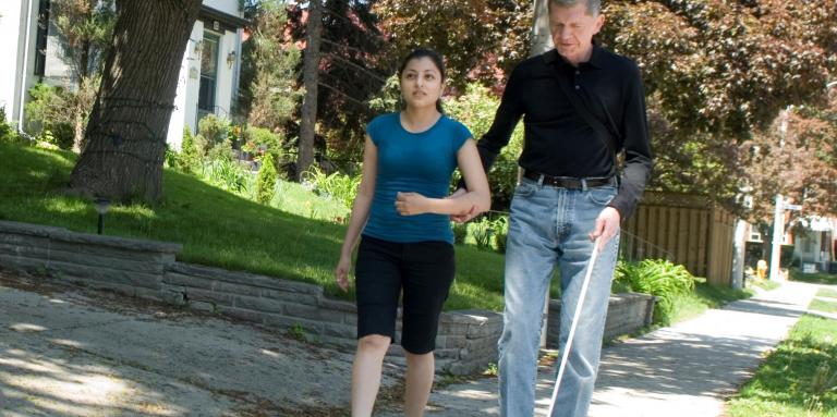 Older man and younger woman walk down sidewalk, man using white cane 