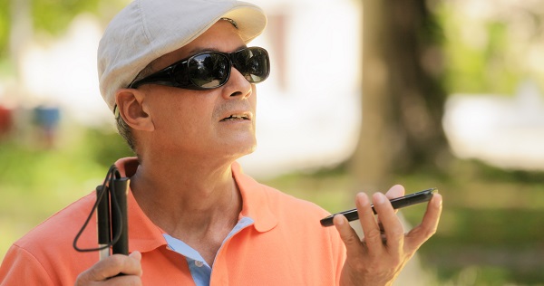 A man wearing dark sunglasses sits in a park, holding a cell phone in one hand and a white cane in the other