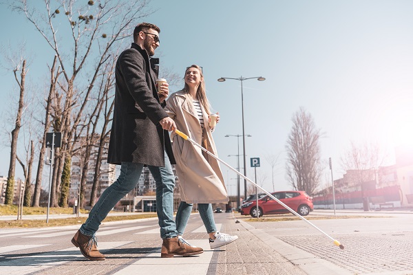 A woman walks across the street with a man who is holding a white cane for navigation