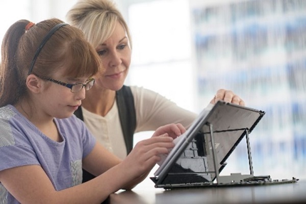 A woman assists a young girl with vision loss as she uses a tabletop device