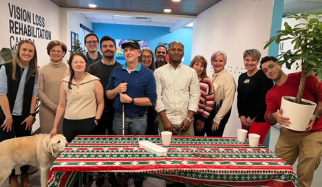 Thirteen people and a Labrador Retriever guide dog gather around a table covered with a festive red, white and green holiday tablecloth.