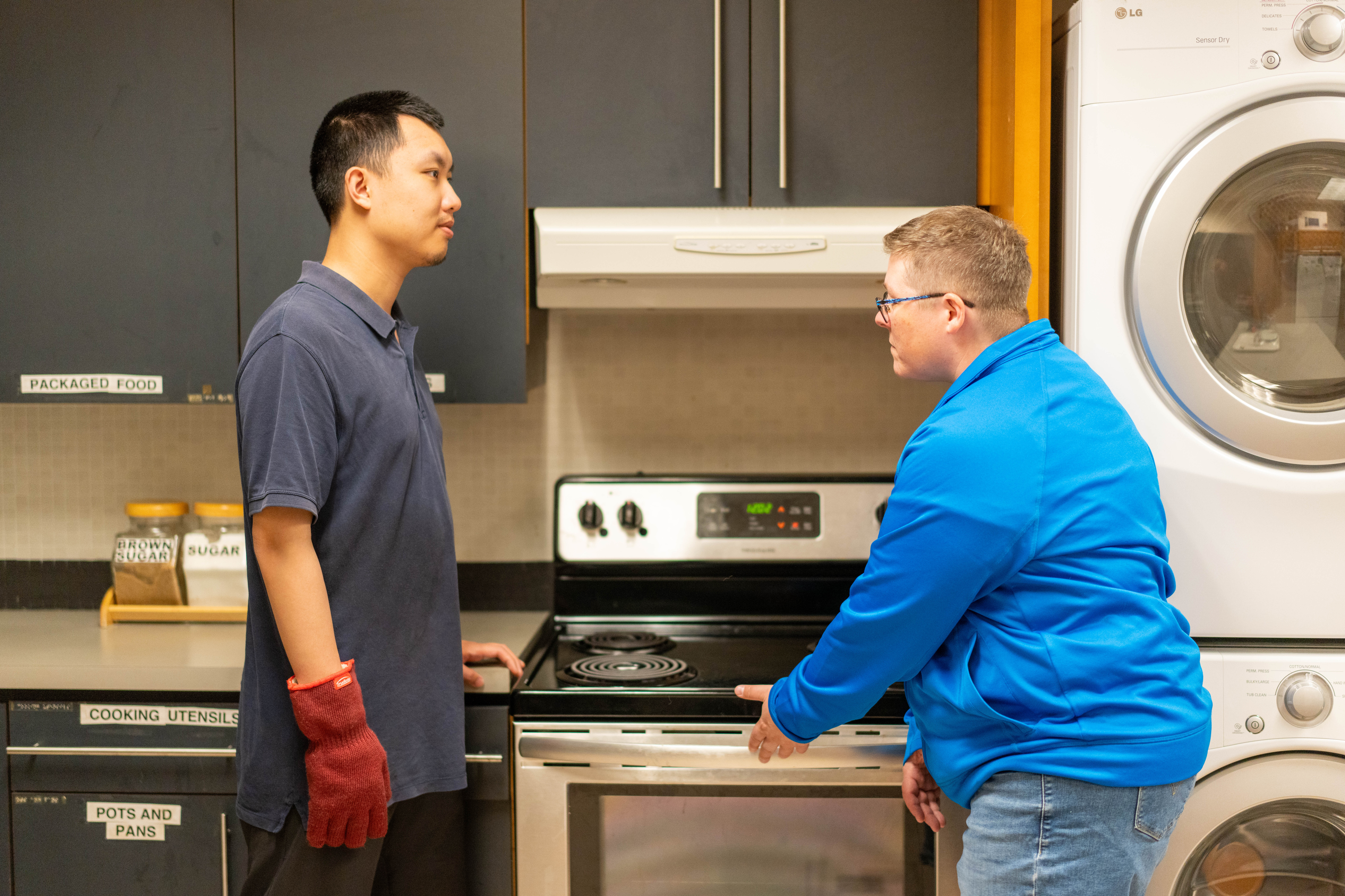 Two individuals are in a kitchen, one in a blue polo shirt and red oven mitt, the other in a blue jacket and glasses. They are standing near a stove and laundry appliances.