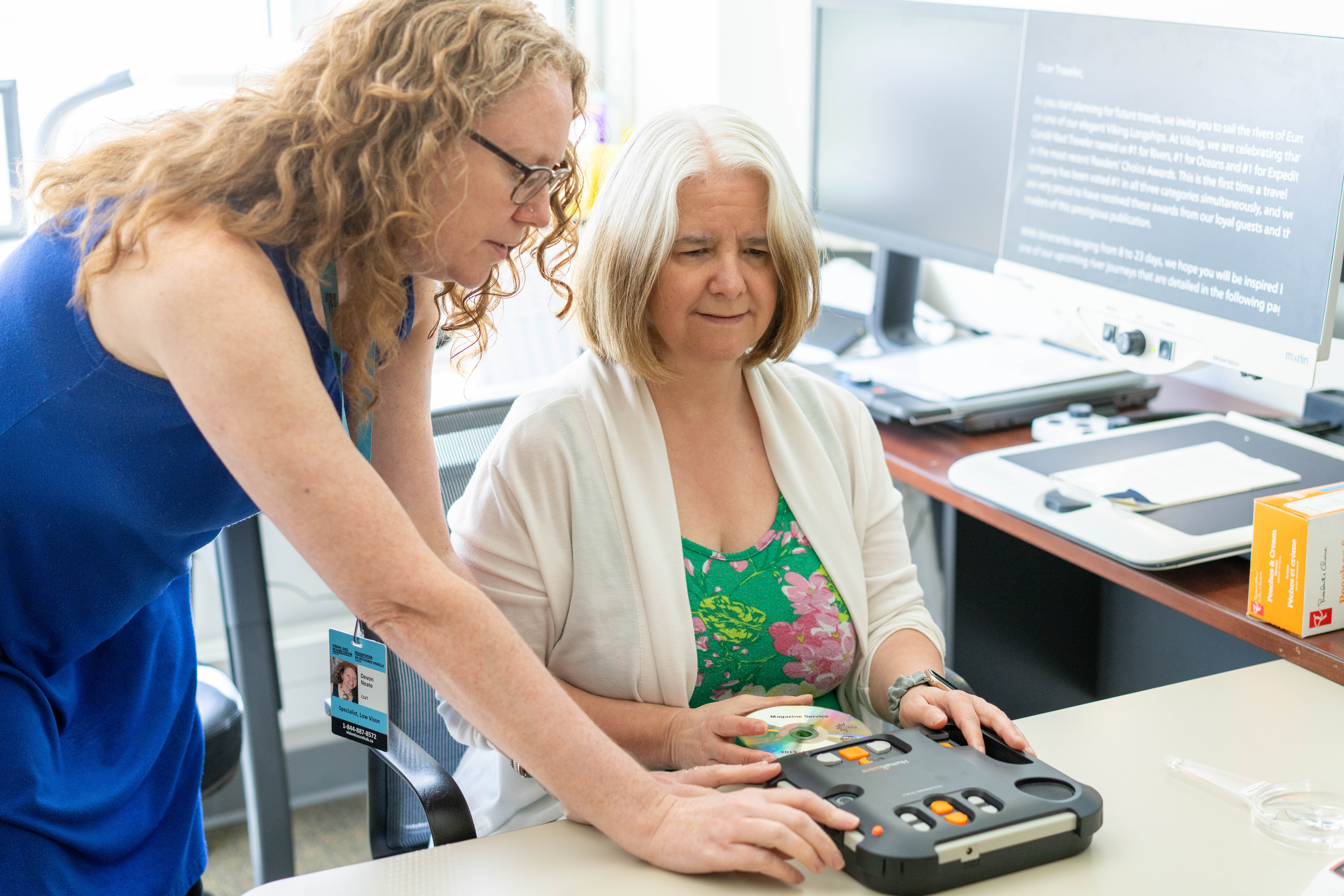 Une femme aux cheveux bouclés se tient à côté d'une femme plus âgée assise à un bureau et suivant une formation sur un appareil tactile, tandis que des écrans d'ordinateur et des technologies d'assistance sont visibles en arrière-plan.