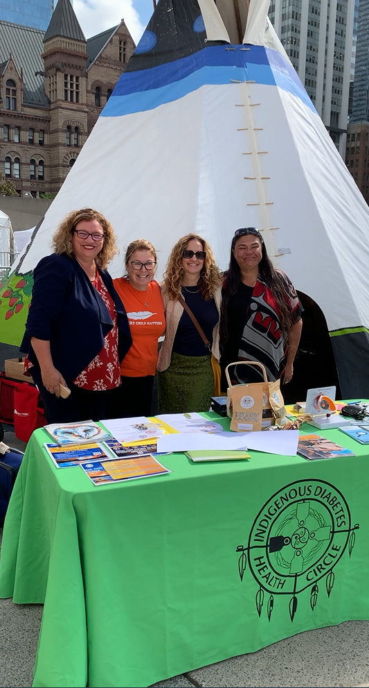 An outdoor outreach table with a green "Indigenous Diabetes Health Circle" tablecloth with 4 women standing behind the table and in front of a teepee. The table displays pamphlets.