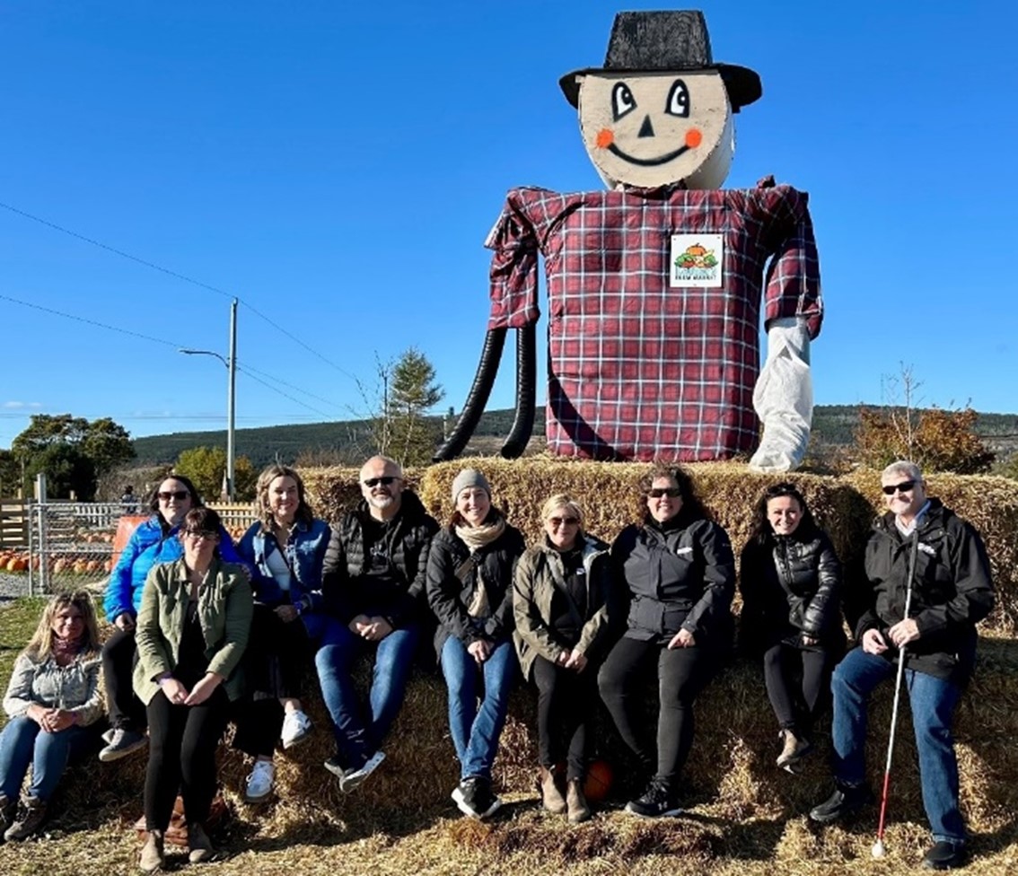 A group of people wearing fall jackets and sitting on bales of hay in front of a giant smiling scarecrow.