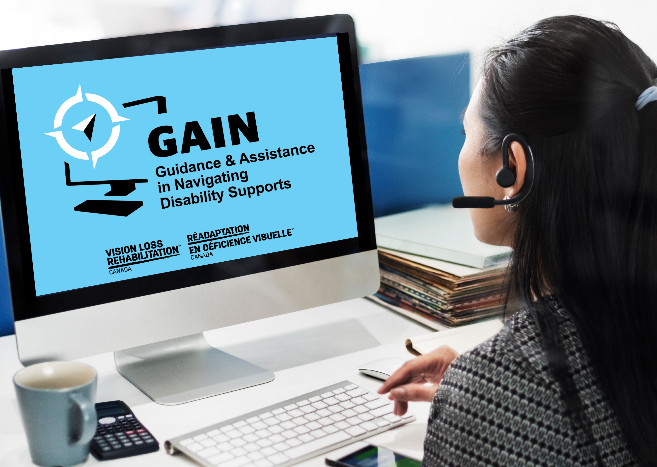 A woman wearing a headset sits at a desk and looks at a computer monitor displaying a blue screen with the logo and text for "GAIN – Guidance & Assistance in Navigating Disability Supports." The screen also shows the logos for "Vision Loss Rehabilitation Canada" and its French equivalent, "Réadaptation en Déficience Visuelle Canada." A keyboard, mug, calculator, and stack of folders are on the desk in front of her.
