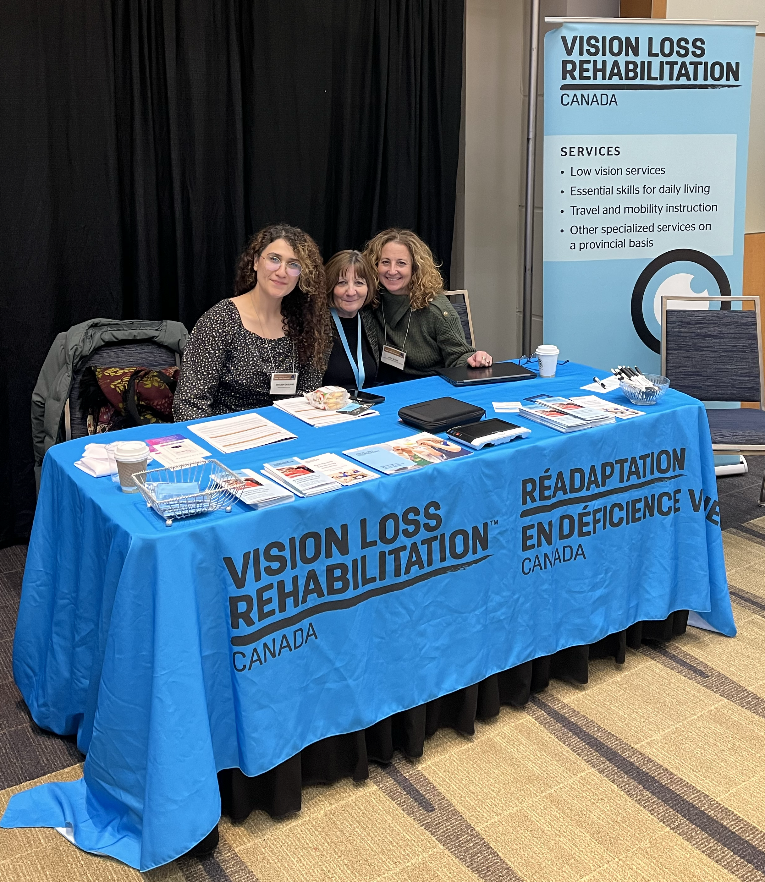 Trois femmes sont assises à une table recouverte d'une nappe bleue dotée du logo RDVC bilingue sur laquelle sont disposés des brochures et des documents, ainsi qu'une bannière présentant les services d'aide pour les personnes ayant une limitation visuelle.