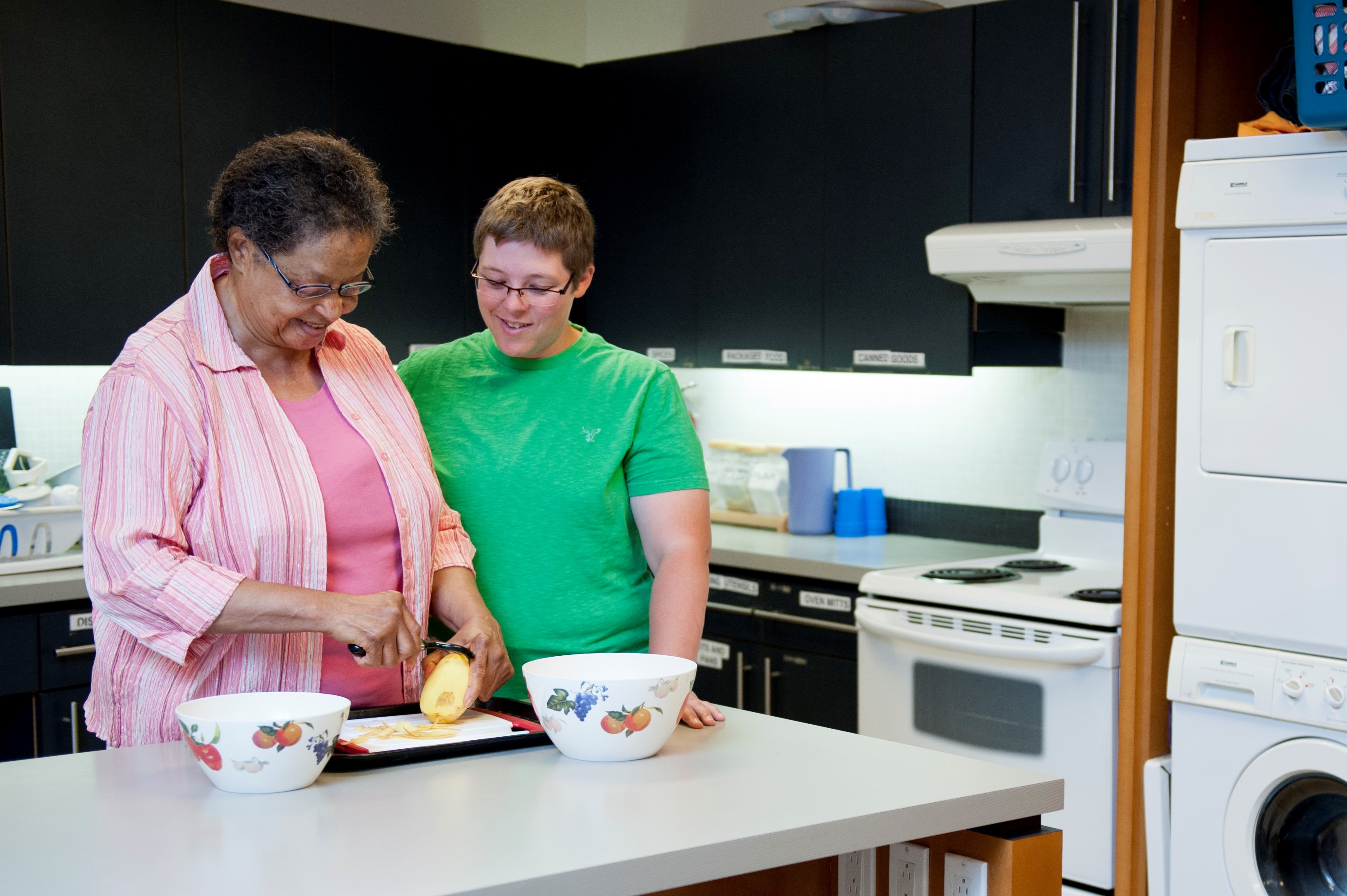 Younger woman watches older woman chop vegetables in kitchen, both smiling 