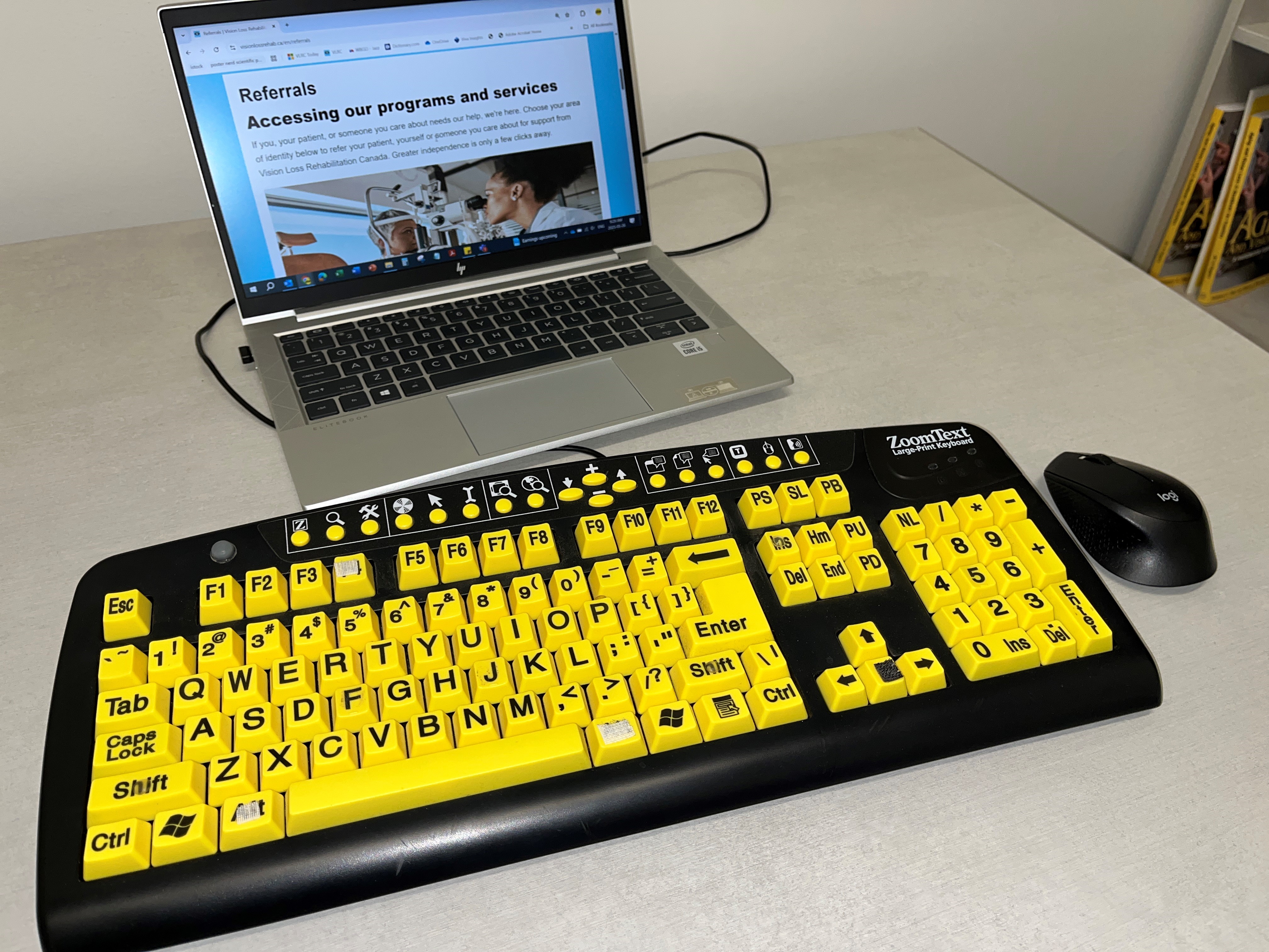 Laptop showing a referrals webpage, with a large-print yellow keyboard and a black mouse on a desk.