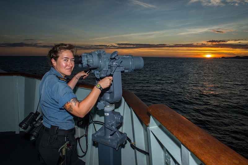 A female Canadian sailor stands on a ship’s deck in front of the deck binoculars (“big eyes”).