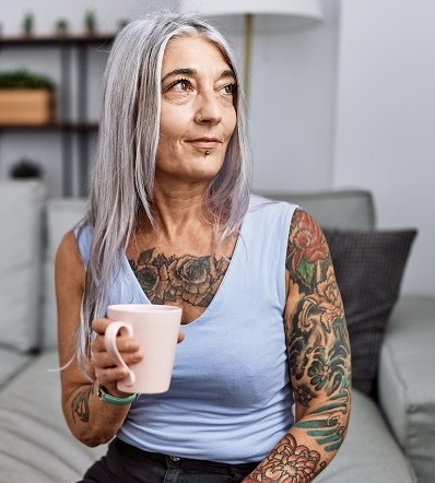 A woman with long grey hair and heavily tattooed sits on a couch and holds a coffee cup