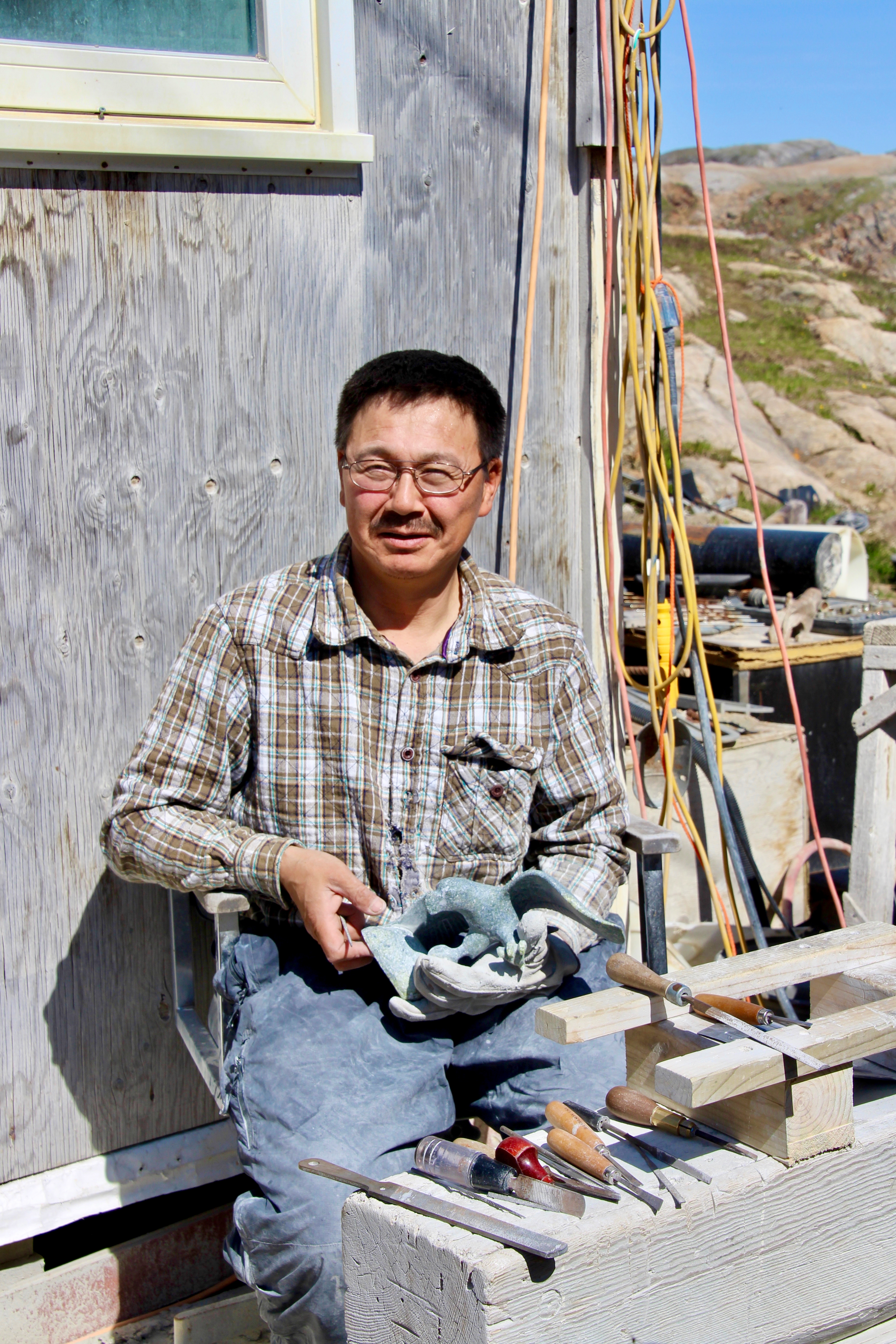 An older Inuit man with glasses is making a soapstone sculpture. He is holding tools and is near a wooden structure. The background shows a rocky landscape.