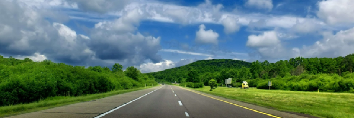 Landscape photo of a highway surrounded by green hills and fields against a blue sky 