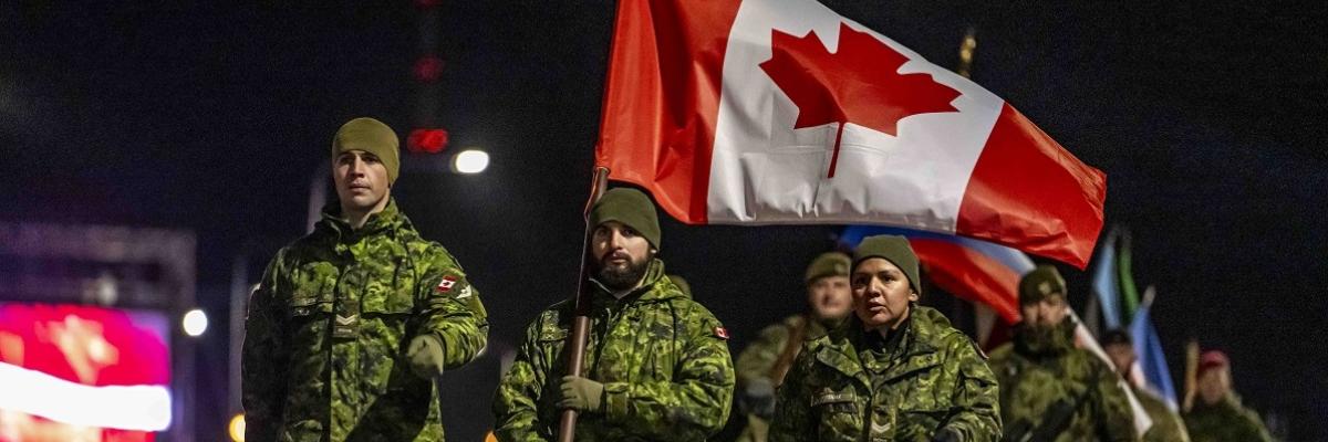 Des soldats canadiens en uniforme de camouflage défilent la nuit, un soldat portant un grand drapeau canadien