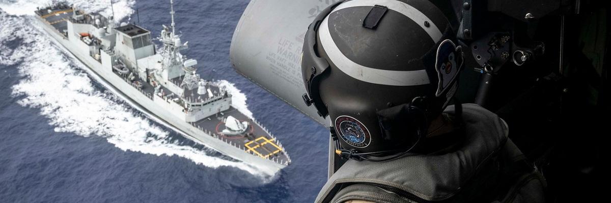 A Canadian soldier sits in the open door of a helicopter, looking down at a naval ship sailing on the ocean.