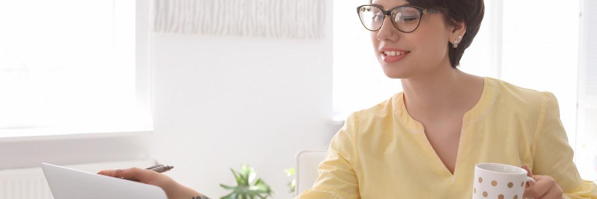 A woman wearing a yellow blouse sits in front of a laptop while holding a coffee cup in her left hand