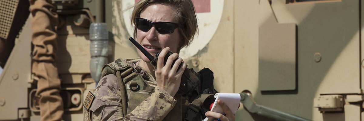 Une femme vêtue d'une tenue de camouflage militaire et portant des lunettes de soleil parle dans un talkie-walkie à côté d'un char d'assaut marqué d'une croix rouge.