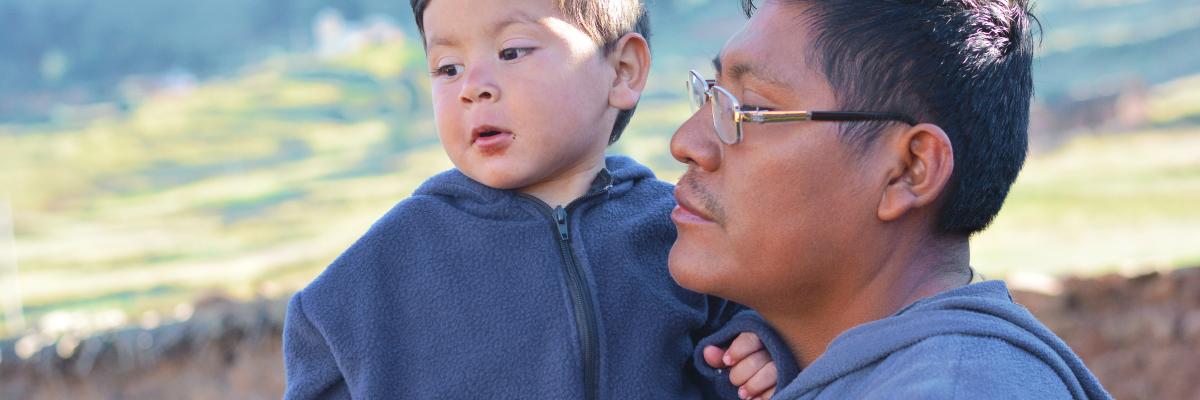 A man with glasses holds a young child in his arms, both wearing dark blue hooded jackets. They are outdoors with a blurred background of hills.