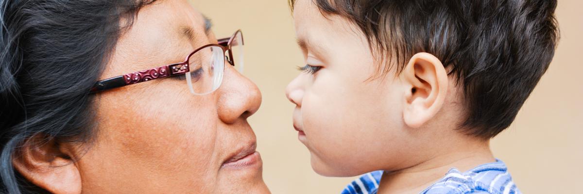 A close-up shot shows an older Indigenous woman with dark hair and glasses looking tenderly at a toddler with dark hair. They are very close to each other, almost touching faces.