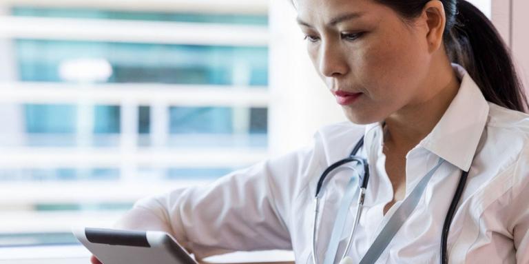 Health care professional looks at tablet, standing by window 