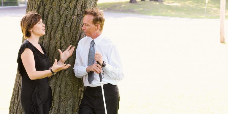 Professional looking man and woman stand by tree talking; man has white cane 