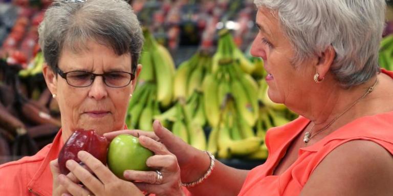 Two woman, wearing coral-coloured tops, are shopping for apples. 