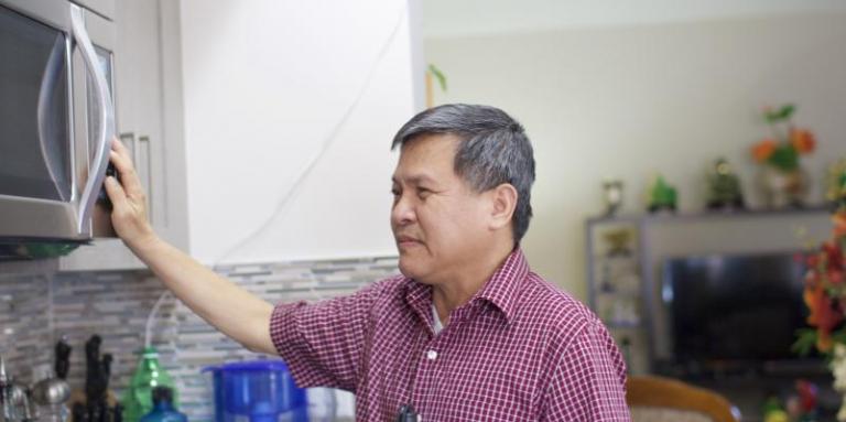 Man in a red plaid shirt is holding his white cane to his side while reaching for the microwave.