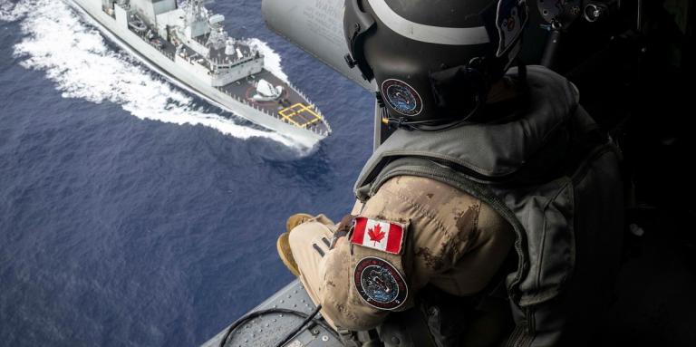 Un soldat canadien est assis dans la porte ouverte d'un hélicoptère et regarde un navire militaire naviguant sur l'océan.