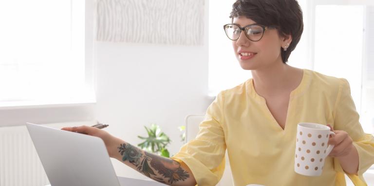 Une femme portant un chemisier jaune est assise devant un ordinateur portable tout en tenant une tasse de café dans sa main gauche