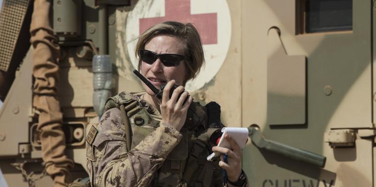 Une femme vêtue d'une tenue de camouflage militaire et portant des lunettes de soleil parle dans un talkie-walkie à côté d'un char d'assaut marqué d'une croix rouge.