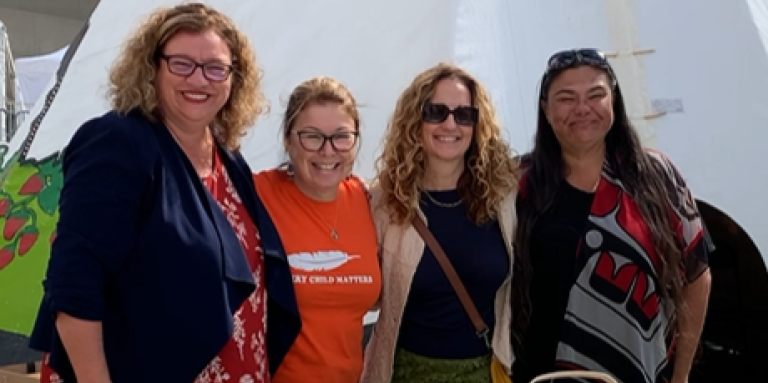 An outdoor outreach table with a green "Indigenous Diabetes Health Circle" tablecloth with 4 women standing behind the table and in front of a teepee. The table displays pamphlets.