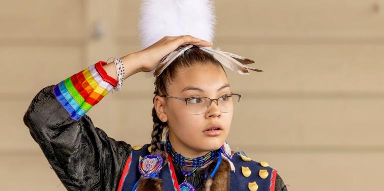 A young Indigenous woman with braids and glasses, wearing traditional clothing with beadwork and a feathered headdress, adjusts the headdress.