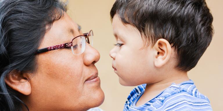 A close-up shot shows an older Indigenous woman with dark hair and glasses looking tenderly at a toddler with dark hair. They are very close to each other, almost touching faces.