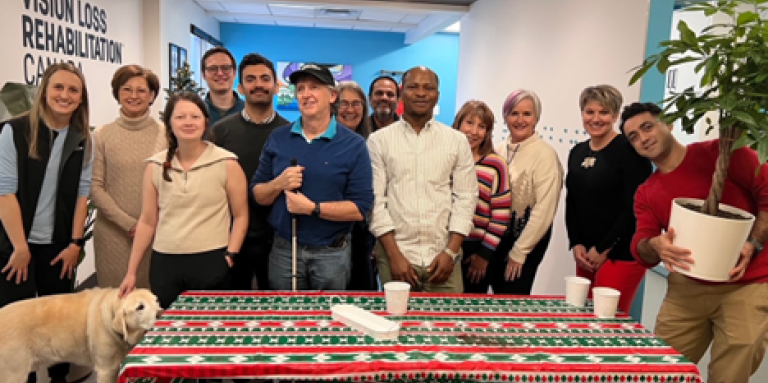 Thirteen people and a Labrador Retriever guide dog gather around a table covered with a festive red, white and green holiday tablecloth.
