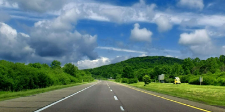 Photographie panoramique d'une autoroute entourée de collines verdoyantes et de champs, avec un ciel bleu en arrière-plan.