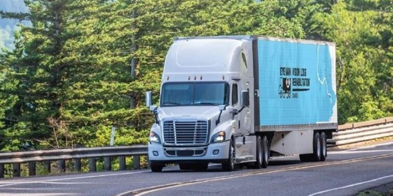 A blue tractor-trailer with the Eye Van logo written on the side, on a curvy highway with evergreen trees on the side of the road.