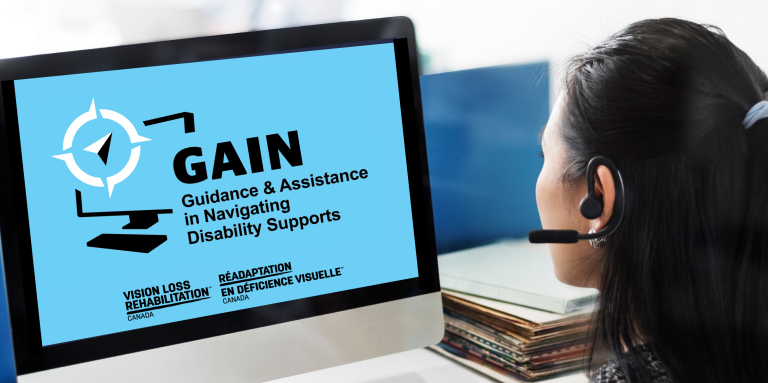 A woman wearing a headset sits at a desk and looks at a computer monitor displaying a blue screen with the logo and text for "GAIN – Guidance & Assistance in Navigating Disability Supports." The screen also shows the logos for "Vision Loss Rehabilitation Canada" and its French equivalent, "Réadaptation en Déficience Visuelle Canada." A keyboard, mug, calculator, and stack of folders are on the desk in front of her.